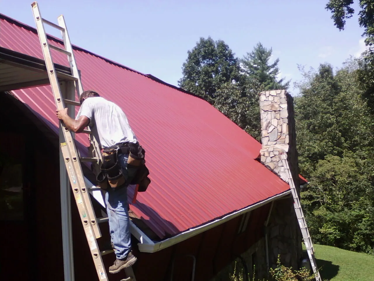 Roofer on a ladder beside a red metal roof and stone chimney — Soffit & Fascia Repair by Roofing Brothers in Homestead